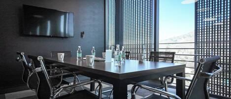 A conference room with a long table, four chairs, a TV mounted on the wall, and a view of the outdoors through large windows.