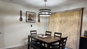 Un coin repas avec une table en bois foncé, quatre chaises, un banc, une suspension, un tableau encadré et une fenêtre avec des stores.