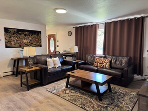A living room with a dark leather sofa, a wooden coffee table, and a patterned rug. There is a painting on the wall and a door with a stained glass window. - Charming 3-BR house in Juneau perfect for your getaway, minutes from the Airport (Juneau)