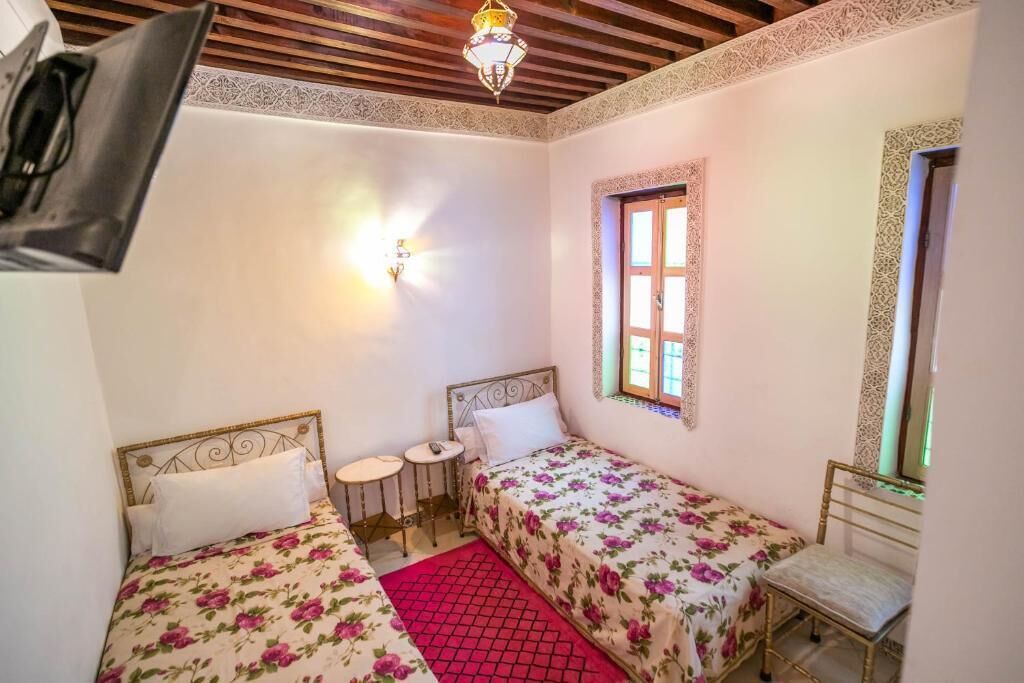 Een slaapkamer met twee enkele bedden, een klein tafeltje, een stoel, een decoratieve spiegel en een raam met versierde luiken.