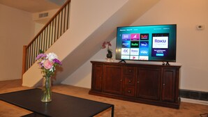 A living room with a flat-screen TV on a wooden stand, a vase with flowers on a table, and a staircase in the background. - Sweet home nearby UOA (Tucson)