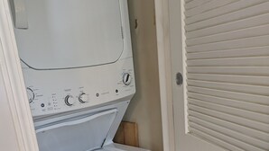 A compact laundry room with a front-loading washer and dryer, a wooden shelf, and a door with a louvered window.