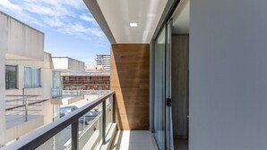 Un balcón con puerta de cristal, barandilla de metal y vista de edificios al fondo.