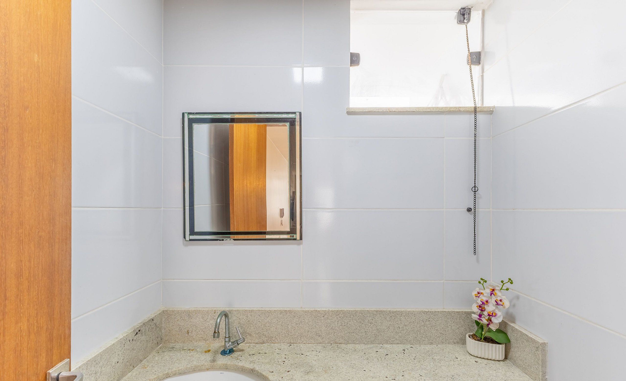 Un baño con azulejos blancos, una puerta de madera, un lavabo con grifo, un espejo, una ducha y una orquídea en maceta.