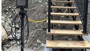 Una escala amb barana metàl·lica negra que condueix a una porta, una caixa elèctrica exterior negra i una unitat de climatització blanca muntada a la paret.