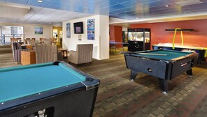 A pool table area with a pool table, a billiards table, and a foosball table. There are chairs and a table with a drink dispenser in the background.