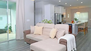 A modern living room with a beige sectional sofa, a dining area with a kitchen in the background, and a sliding glass door leading to a balcony.