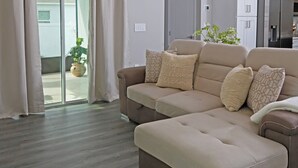 A modern living room with a sectional sofa, a pendant light, and a sliding glass door leading to a balcony with a potted plant.