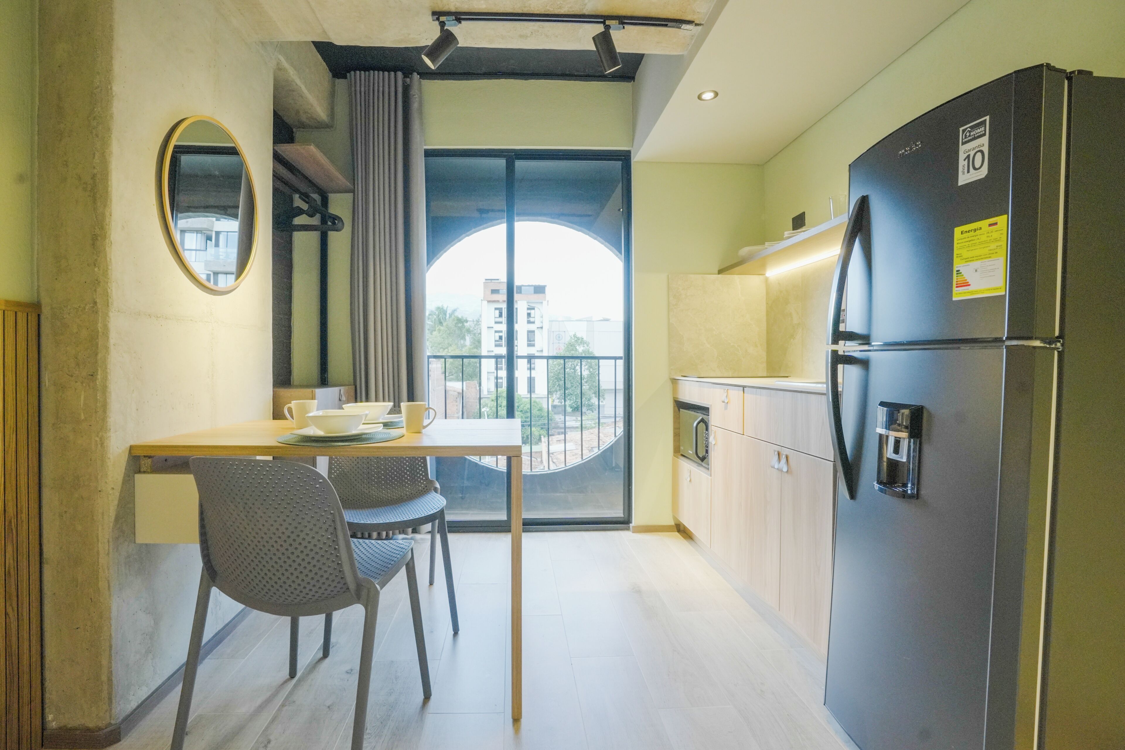 A compact kitchen with a refrigerator, a dining table with chairs, a window with curtains, and a balcony with a view.