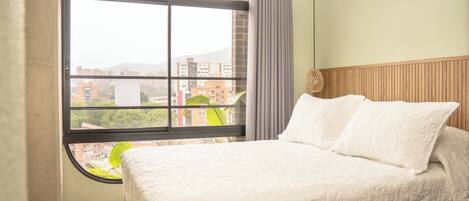 A hotel room with a large window, a bed with white bedding, and a wooden headboard.