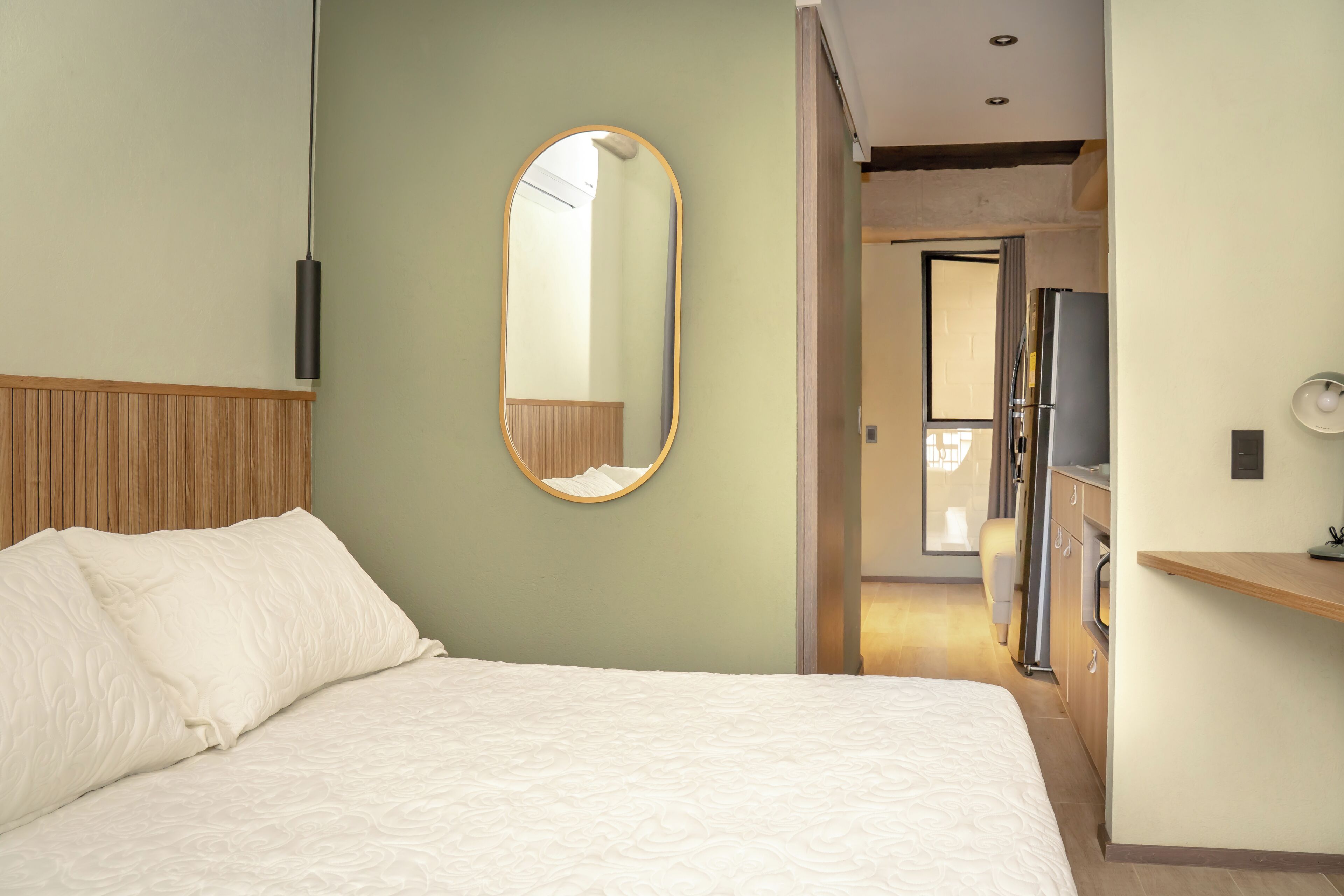 A hotel room with a bed, a mirror, a wooden headboard, a kitchenette, and a door leading to another room.