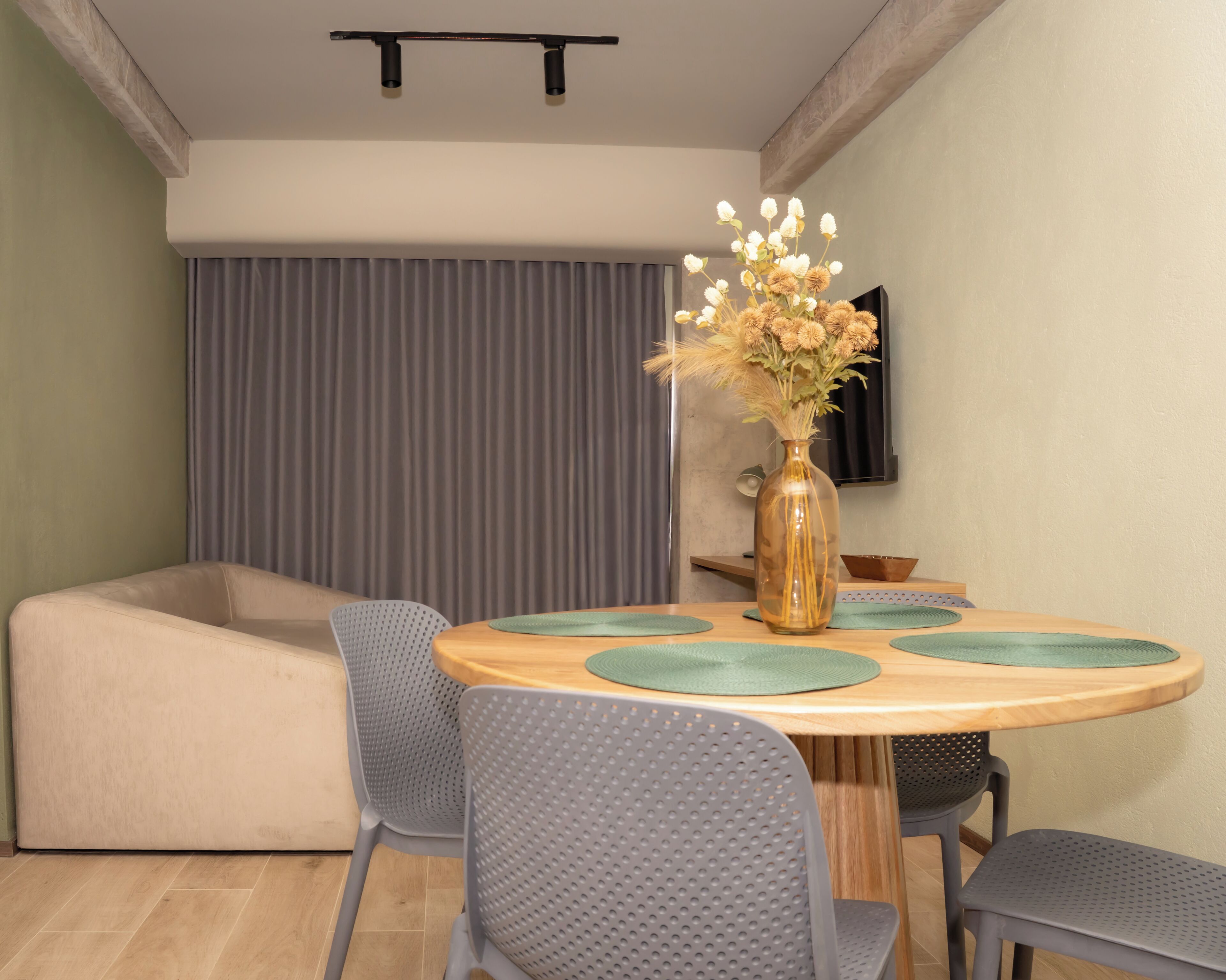 Un área de comedor con una mesa redonda de madera, sillas grises perforadas, un sofá color beige, un florero con flores y un televisor montado en la pared.