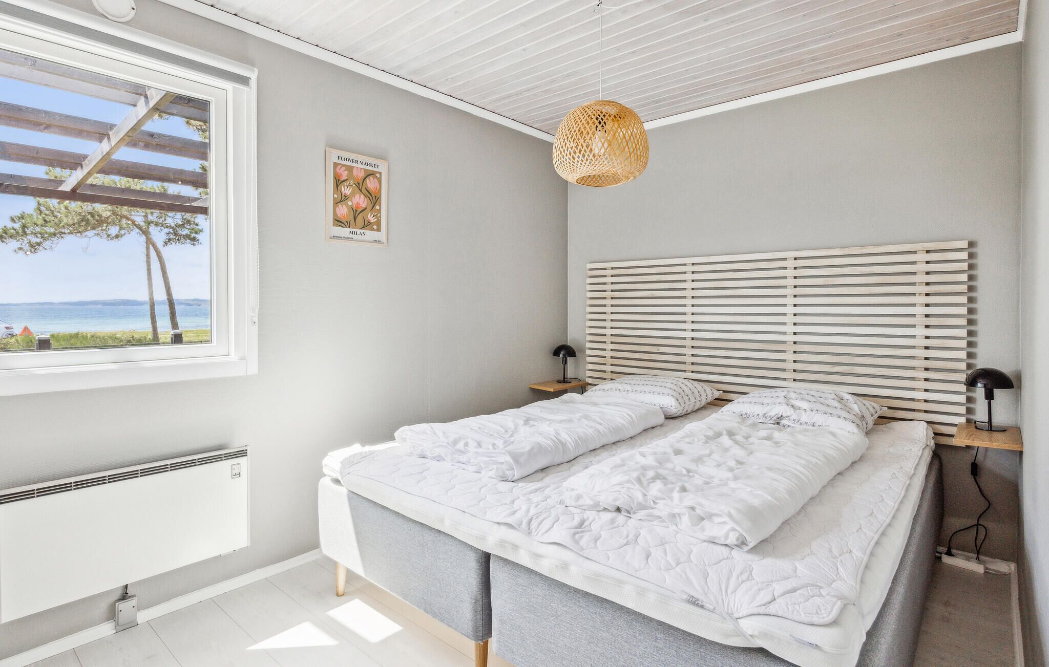 Un dormitorio con cama, ventana con vista al mar, lámpara de pared e imagen enmarcada.