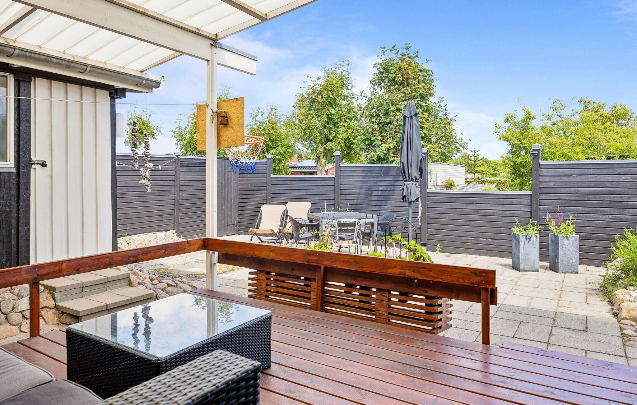 Un patio exterior con terraza de madera, una mesa de vidrio, sillas de mimbre y una parrilla.