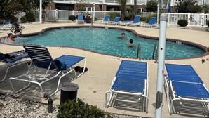 Una piscina con sillones de descanso, un edificio con balcones y un cielo despejado.