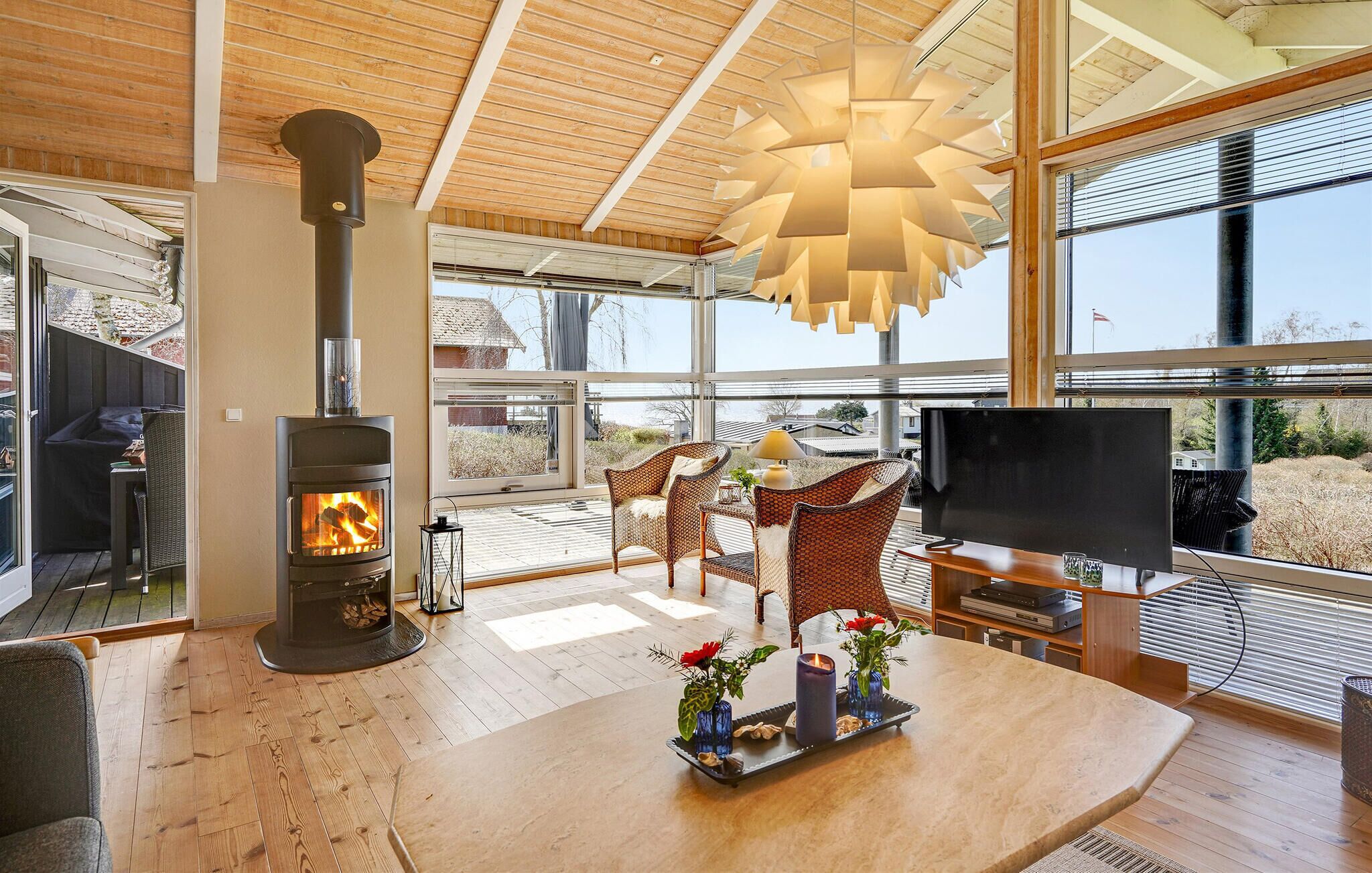 Ein modernes Wohnzimmer mit Kamin, Korbmöbeln, einem Flachbildfernseher und großen Fenstern mit Blick ins Freie.