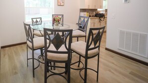 Een eetruimte met een ronde glazen tafel, omringd door vier stoelen, een kroonluchter en een raam met jaloezieën.