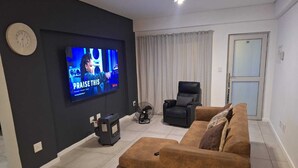 Un salon équipé d’un grand téléviseur à écran plat, d’un fauteuil en cuir noir, d’un canapé marron et d’un ventilateur sur pied.