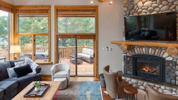 A cozy living room with a stone fireplace, large windows, and a TV mounted above it. - Mountain home w/ AC, BBQ & patio (Whistler)