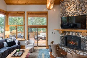 A cozy living room with a stone fireplace, large windows, and a TV mounted above it. - Mountain home w/ chef's kitchen & fireplace (Whistler)