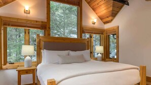 A wooden-framed bedroom with a large bed, two bedside tables with lamps, and windows offering a view of trees. - Mountain home w/ AC, BBQ & patio (Whistler)
