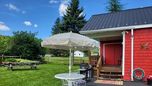 Un patio con una mesa, sillas y una sombrilla, y un edificio rojo con una terraza de madera.