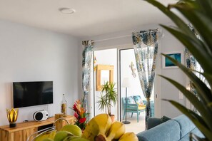A modern living room with a flat-screen TV, a wooden side table, a radio, and a variety of plants. - Odyssée 5 stars, 2 suites, bay view, ODYSSÉE, Votre Parenthèse Céleste (Les Trois-Îlets)