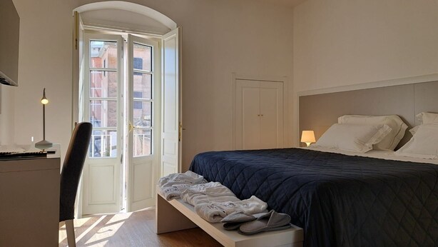 Un dormitorio con cama, escritorio y silla. Se ve una ventana con vistas a edificios.