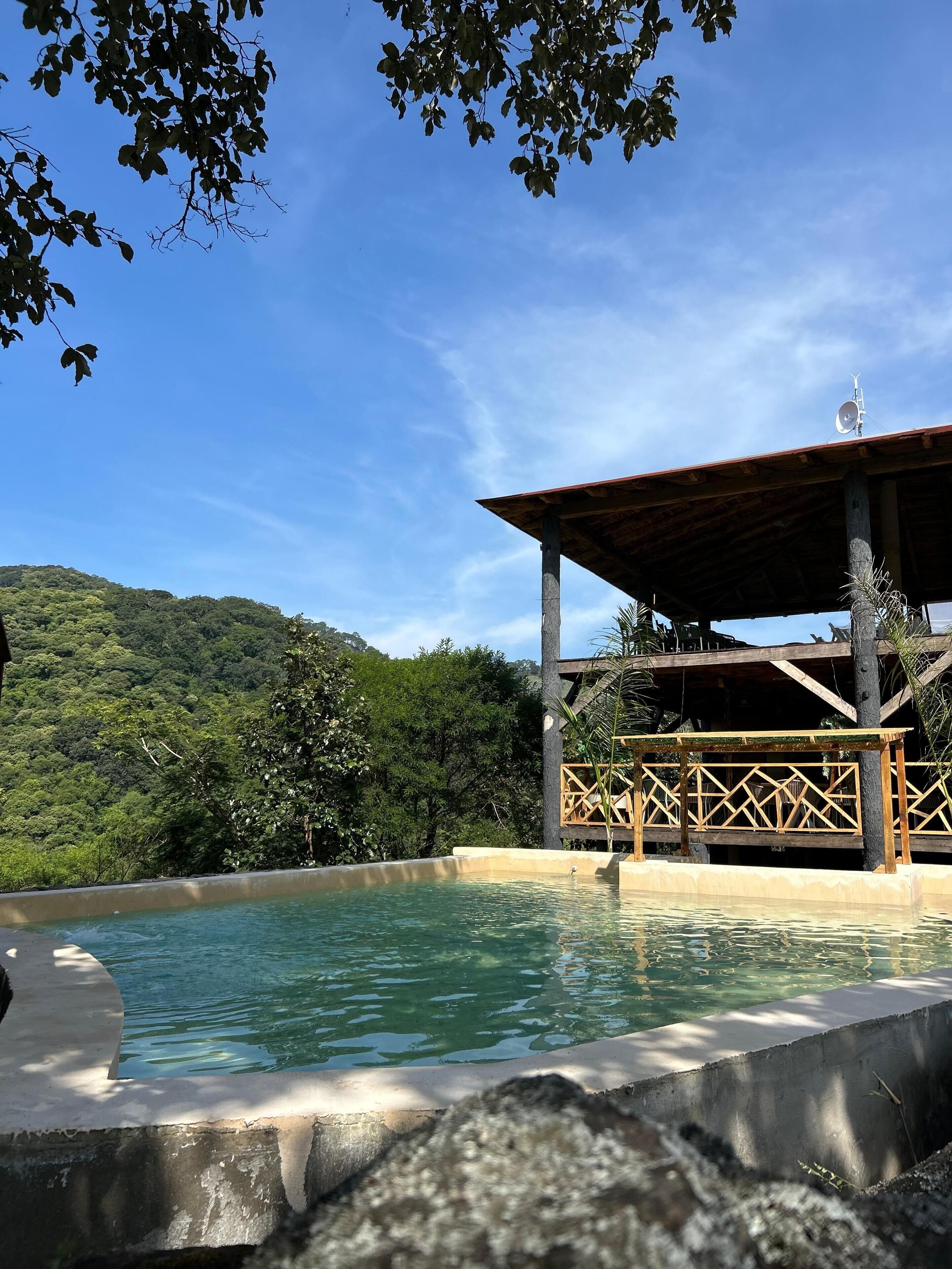Área de piscina com deck de madeira e vista para um morro verdejante.