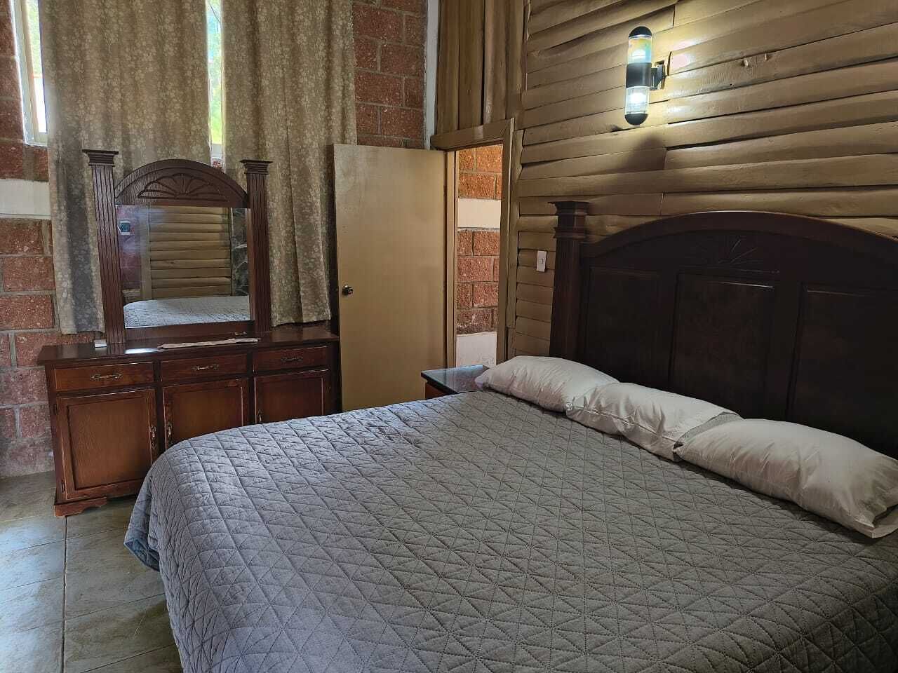 Un dormitorio con cabecera de madera, cama con cubrecama acolchado, cómoda con espejo y lámpara fijada a la pared.