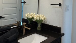 A bathroom with a black countertop, a white sink, a mirror, and a vase with white flowers.