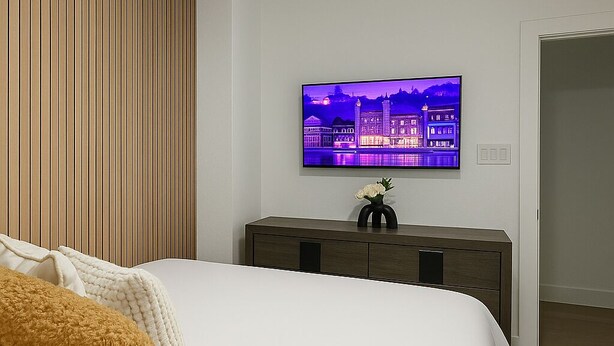 A modern bedroom with a large bed, a wooden panel wall, a TV displaying a cityscape, and a nightstand with a vase.