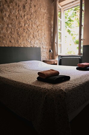 A room with a stone wall, a bed with a patterned bedspread, a window with a view of greenery, and a radiator. - Le Mas de la Digue - Provence (Châteaurenard)