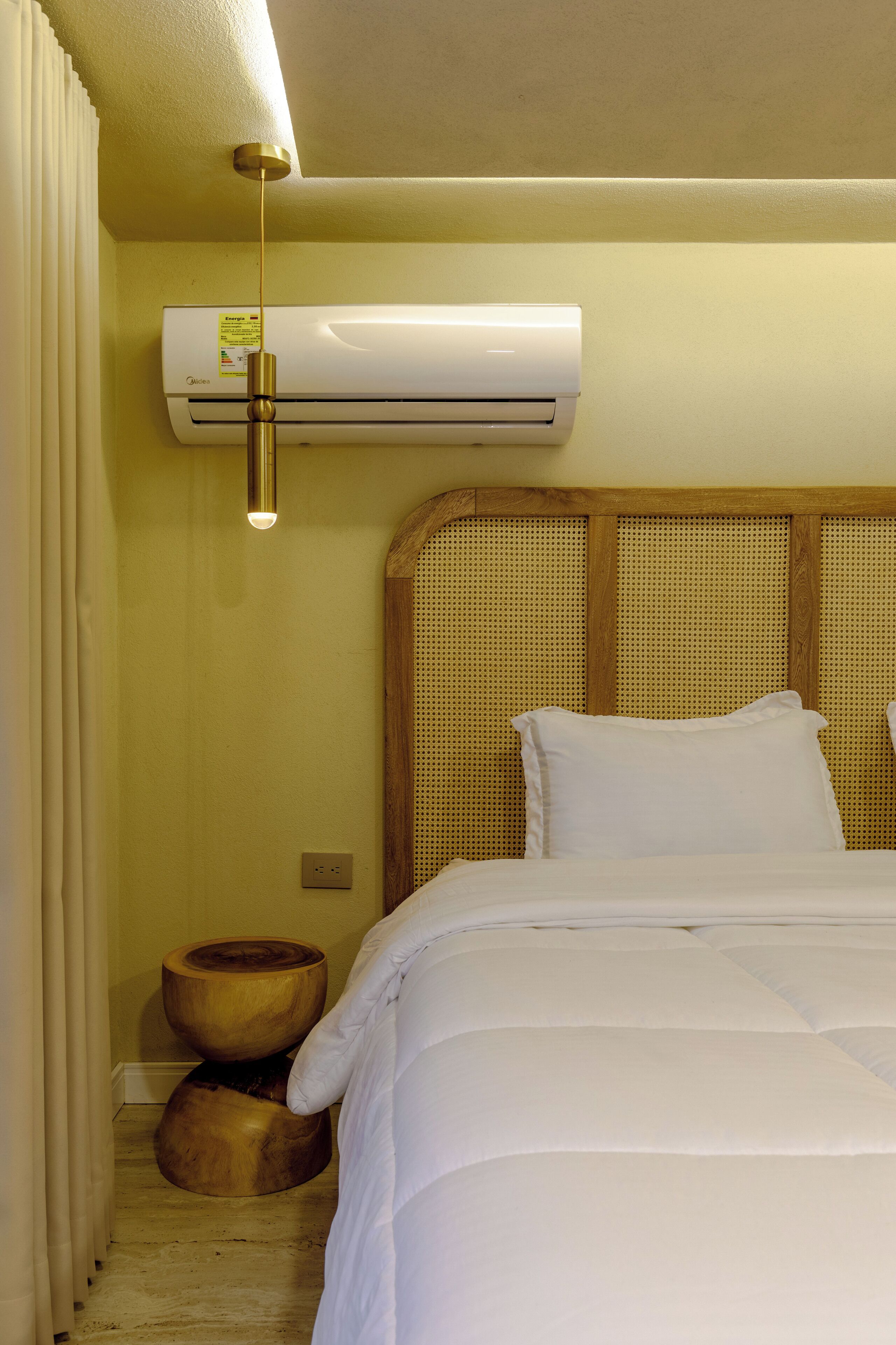 A hotel room with a bed, an air conditioning unit, a wooden headboard, and a small wooden stool.