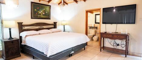 Un dormitorio con techo de madera, una cama con cabecera, una mesita de noche con lámpara, un aparador, un televisor y un cuadro en la pared.