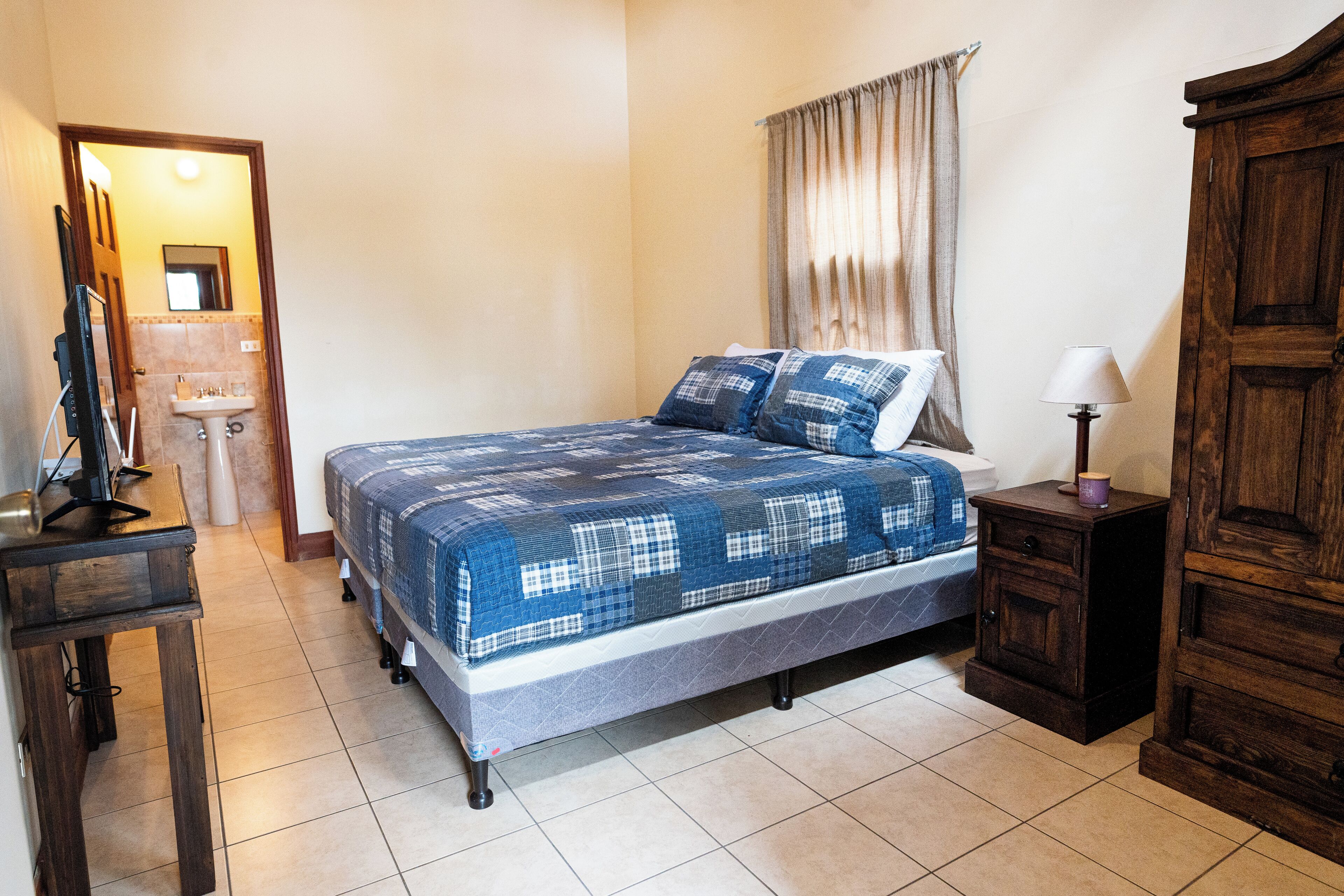 A bedroom with a bed, a bedside table, a lamp, a wardrobe, and a bathroom with a sink and mirror.