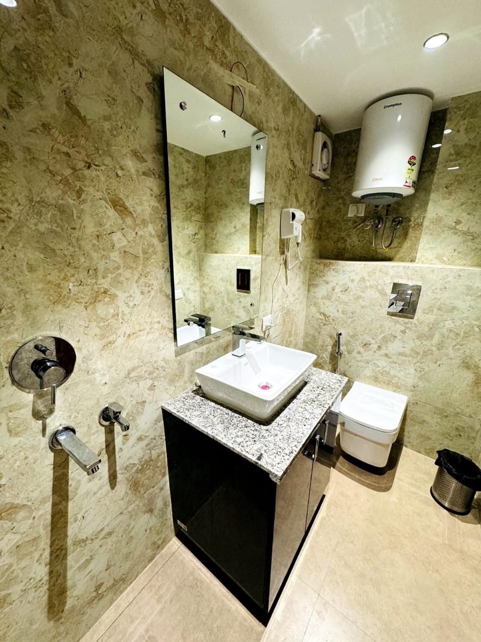 A bathroom with a marble wall, a sink, a toilet, a mirror, and a hand dryer.