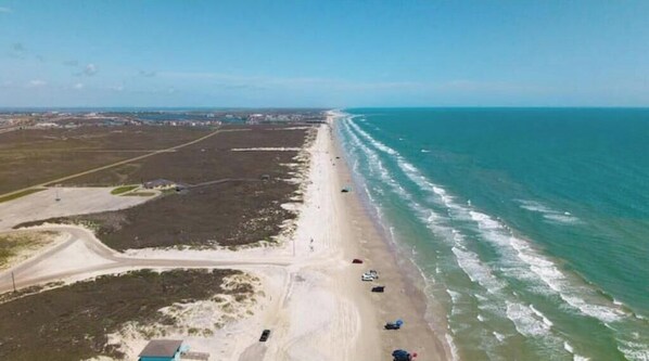 Vista aérea de una playa con una carretera que corre paralela a la orilla, algunos vehículos estacionados a lo largo de la playa, y un cielo despejado.