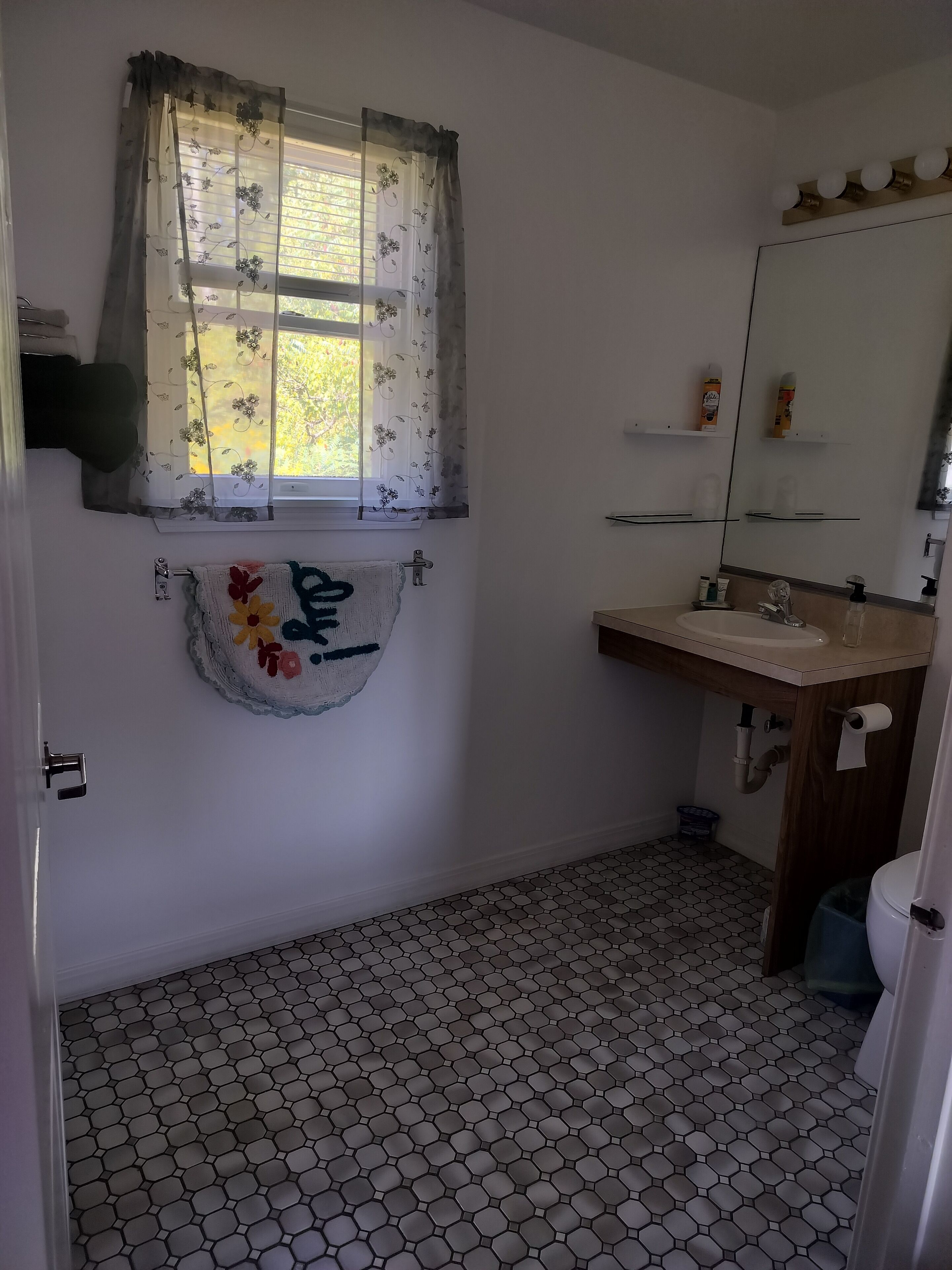 Une salle de bain avec un sol carrelé à motifs hexagonaux, une fenêtre ornée de rideaux à fleurs, une serviette aux couleurs vives, un lavabo avec un miroir et des toilettes.