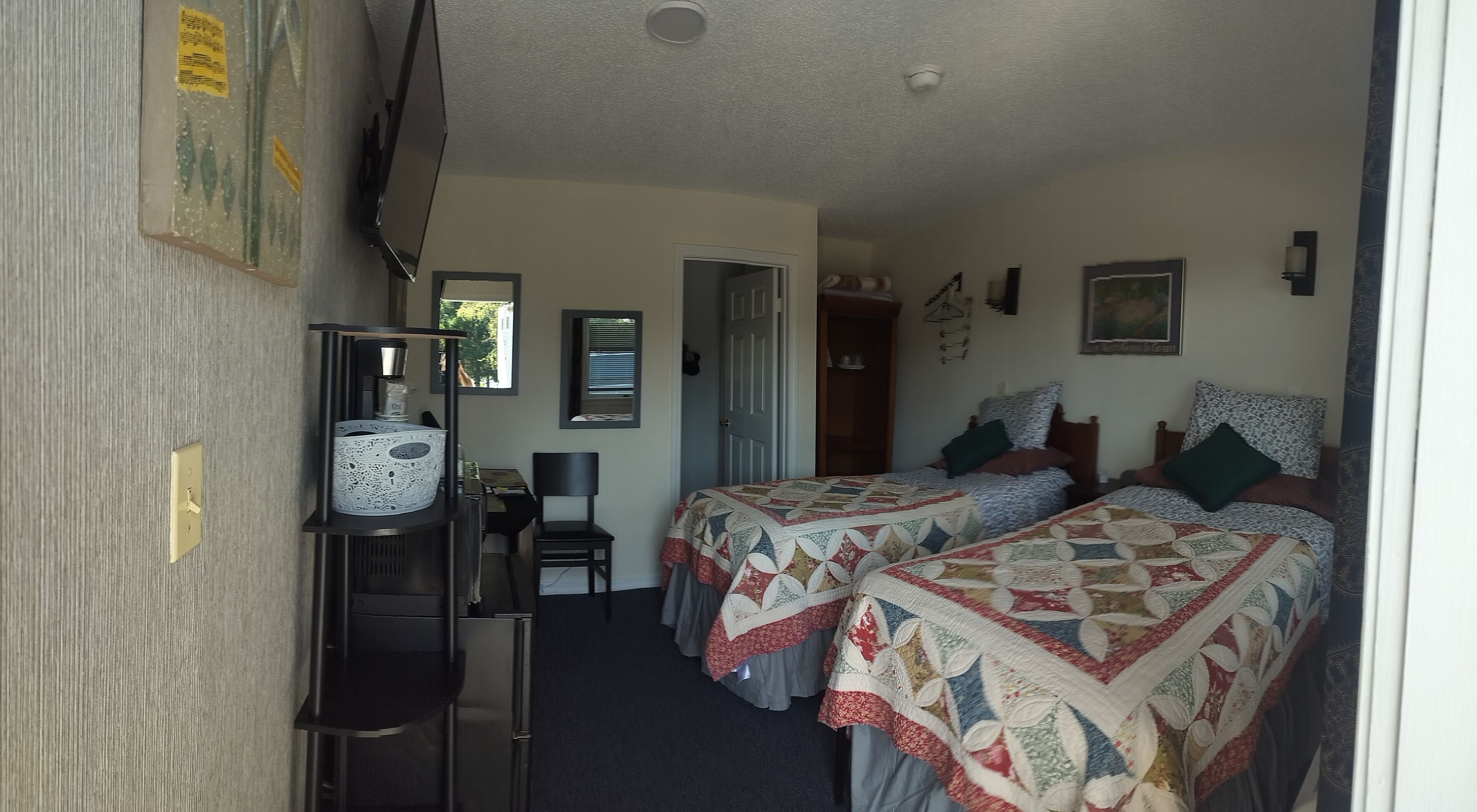 A room with two beds, a desk, a chair, a shelf with a kettle, and a door leading to another room.