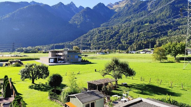 A modern two-story building with a flat roof, surrounded by a well-maintained green field and a mountain backdrop. - 85 m2 Ferienwohnung mit überdachtem Parkplatz und Außenstellplatz - Bergblick (Bürs)