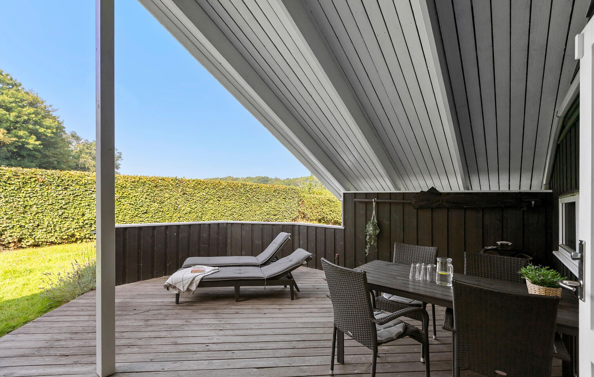 A covered outdoor area with a wooden deck, two lounge chairs, a table with chairs, and a small grill.