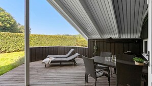 A covered outdoor area with a wooden deck, two lounge chairs, a table with chairs, and a small grill.
