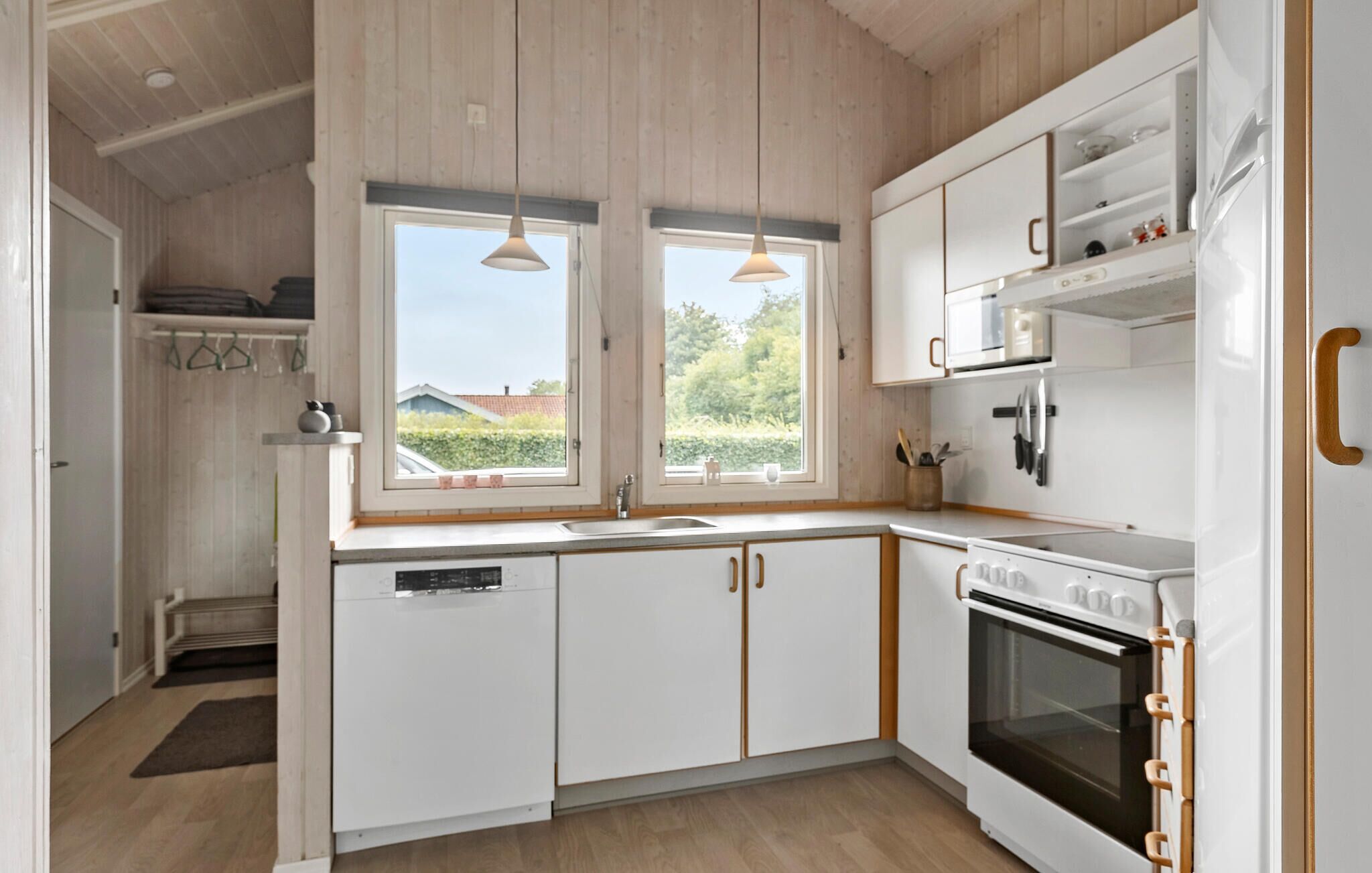 Une cuisine moderne avec des meubles blancs, un lave-vaisselle, une cuisinière et un four à micro-ondes. Deux fenêtres laissent entrer la lumière naturelle et offrent une vue sur l’extérieur.
