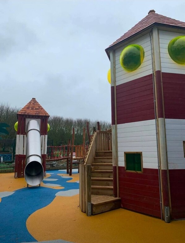A playground with a slide, climbing area, and a building with windows and a door. - Sand Le Mere  (Hull)