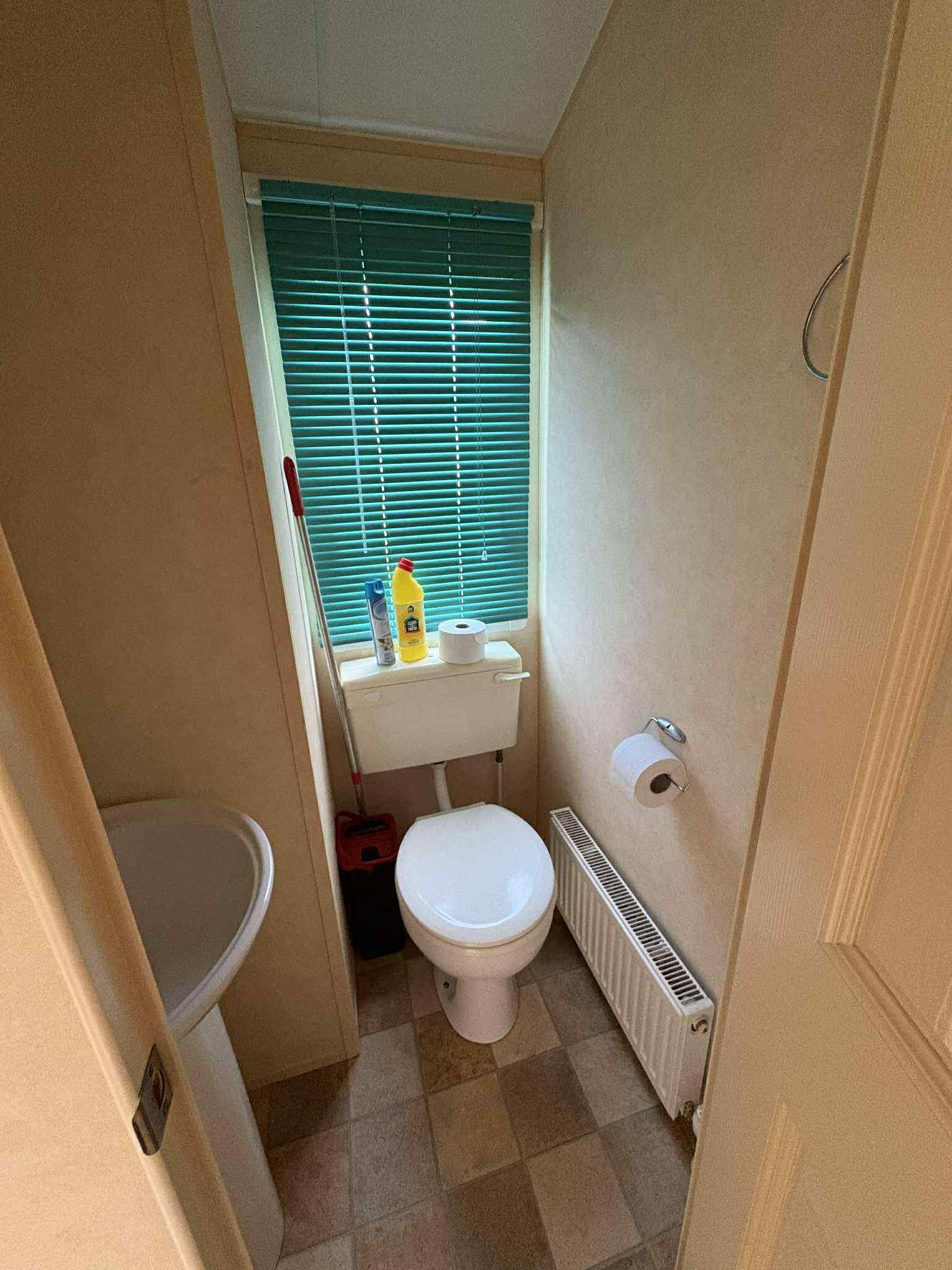 A small, compact bathroom with a toilet, a window with blinds, a radiator, and a small shelf with cleaning supplies.