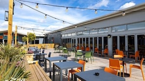 An outdoor patio area with tables and chairs, string lights, and a building with large windows in the background.