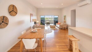 A modern living room with a wooden dining table, white chairs, and a sofa. There are woven wall decorations and a large window with a view. - Luka on Town View Terrace (Margaret River)