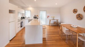 A modern kitchen with white cabinets, a central island, and a dining area with a wooden table and chairs. - Luka on Town View Terrace (Margaret River)
