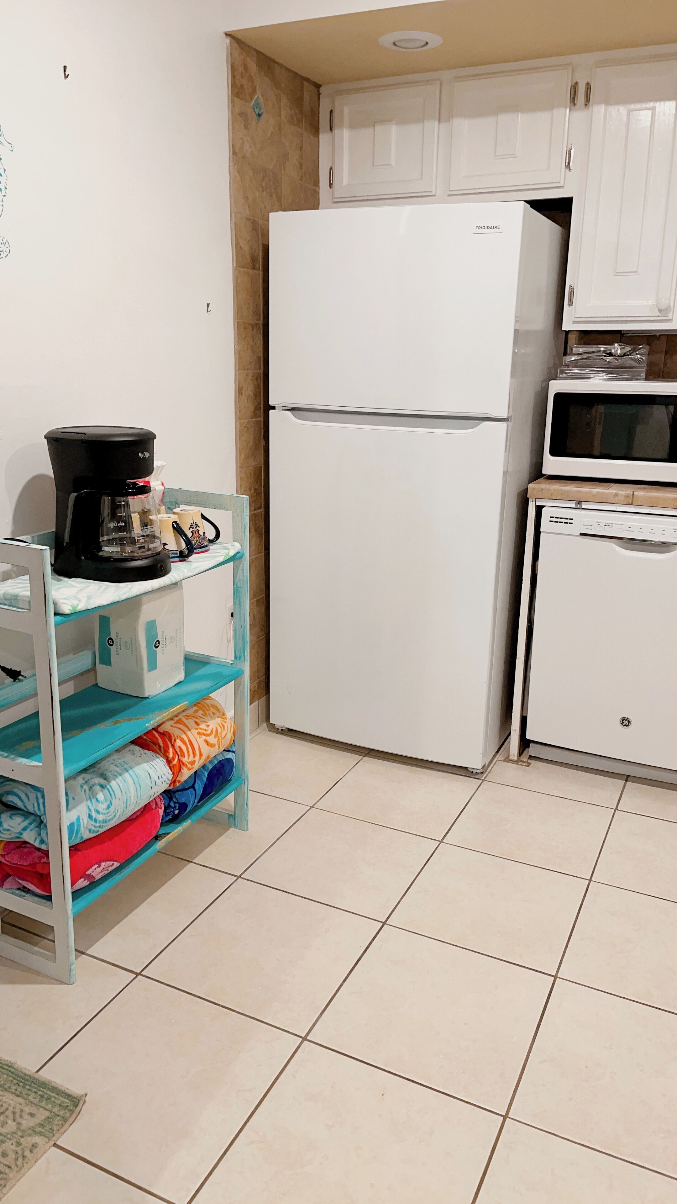 Una cocina con armarios blancos, un refrigerador blanco, un microondas y una repisa con mantas dobladas.
